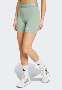 Green Techfit sports shorts made of stretchy material, featuring a wide waistband and white Adidas logo on the left leg. White athletic shoes and socks.