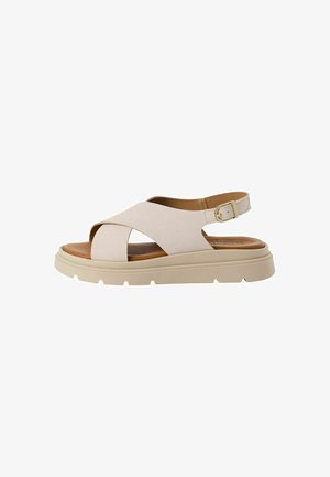 Beige sandal med krydsbåndsdesign, justerbar ankelrem, polstret fodseng og tyk, struktureret sål. Accentueret med en guldspænde.
