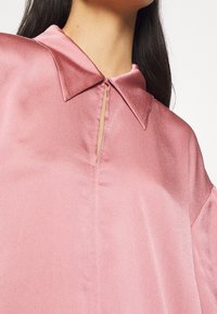 Silky, light pink blouse with a small collar, keyhole detail, and slightly puffed sleeves. Smooth texture with subtle sheen.