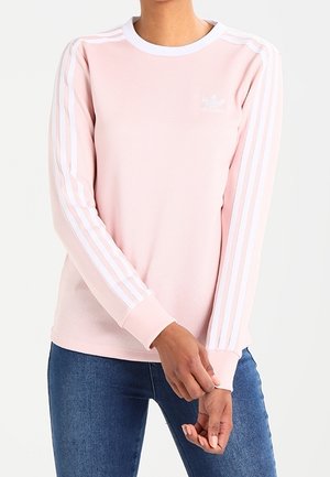 Personne portant un t-shirt Adidas rose pâle à manches longues avec des rayures blanches sur les manches et un jean bleu, mains ajustant le poignet.