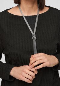 Zilveren metalen ketting met een geknoopt hanger, gedragen over een textuur zwart top met een brede halslijn en lange mouwen.