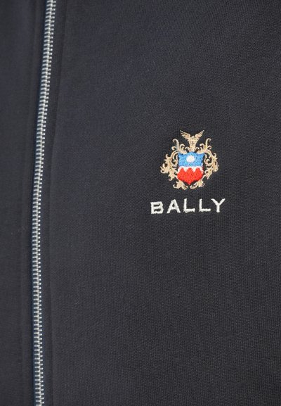 Chaqueta navy con cremallera, de textura suave, que presenta un emblema bordado con acentos en rojo y azul y el texto "BALLY" en blanco.