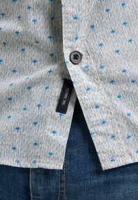 Camicia azzurro chiaro a fantasia con piccoli motivi blu scuro, dotata di chiusura a bottoni e di un'etichetta laterale, abbinata a jeans in denim.