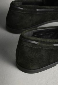 Gros plan sur des mocassins en daim vert foncé montrant l'intérieur en cuir texturé et la semelle en caoutchouc lisse sur une surface gris clair.