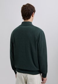 Maglione verde scuro realizzato in tessuto morbido, caratterizzato da collo alto e maniche lunghe. Texture liscia con polsini e orlo a coste.