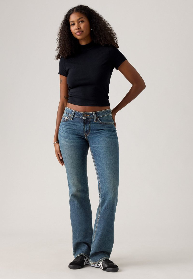 Black cropped t-shirt with short sleeves, paired with mid-blue flared jeans featuring a button fly and standard five-pocket design. Black sneakers.