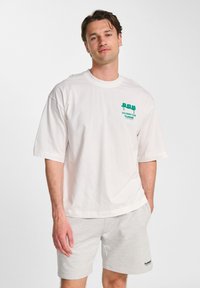 White cotton T-shirt with short sleeves featuring a green graphic design on the left chest, paired with light gray shorts.