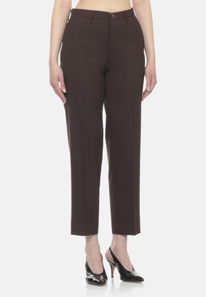 Donna che indossa pantaloni dritti marrone scuro corti e décolleté nere con cinturino, in piedi davanti a uno sfondo semplice.