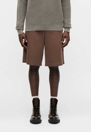 Oseba, oblečena v rjave hlače do kolen, bež pleten pulover, bele nogavice in temno rjave vezalne škornje na enostavni ozadju.