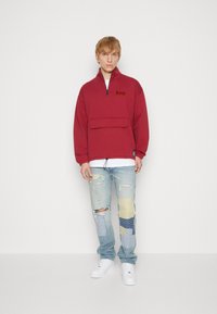 Pullover rosso con colletto alto e tasca frontale, abbinato a jeans chiari con design a toppe nei toni blu e giallo. Sneakers bianche.