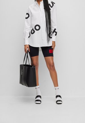 Femme portant une chemise blanche oversize avec des lettres noires, un short de cyclisme noir avec un patch rouge, des chaussettes blanches avec des sandales noires, et tenant un sac fourre-tout noir.