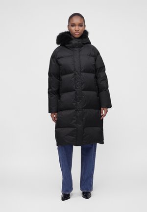 Femme debout portant un long manteau noir matelassé avec une capuche bordée de fourrure, un jean bleu et des chaussures noires pointues sur un fond uni.