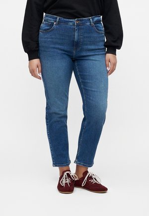 Persona che indossa jeans blu skinny, una maglietta nera a maniche lunghe e sneakers rosse con lacci bianchi, in piedi su uno sfondo semplice.