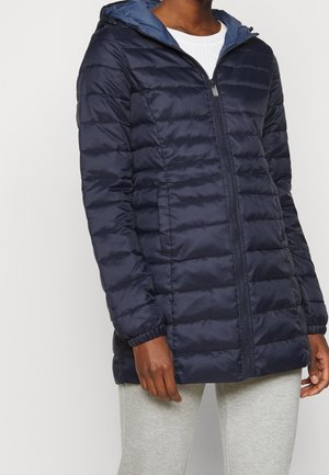 Personne portant une veste matelassée bleu marine à capuche avec fermeture éclair à l'avant, poignets élastiques, sur un haut blanc et un pantalon gris clair.