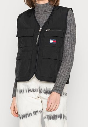 Zwarte Tommy Jeans bodywarmer met meerdere zakken, gedragen over een grijs geribbelde coltrui en witte tie-dye broek.
