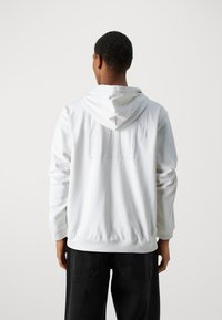 Sudadera blanca con cordón, que presenta un logo en relieve en la parte posterior. Hecha de tela suave, tiene mangas largas y un corte relajado.