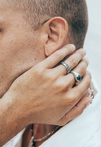 Silverringar med intrikata mönster, en med en grön sten, som vilar på en hand nära en närbild av ett ansikte med kort hår.