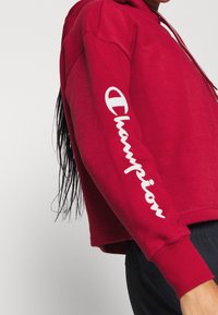 Czerwona bluza z kapturem o krótkim kroju, z białym logo "Champion" na lewym rękawie i teksturowanym materiałem.