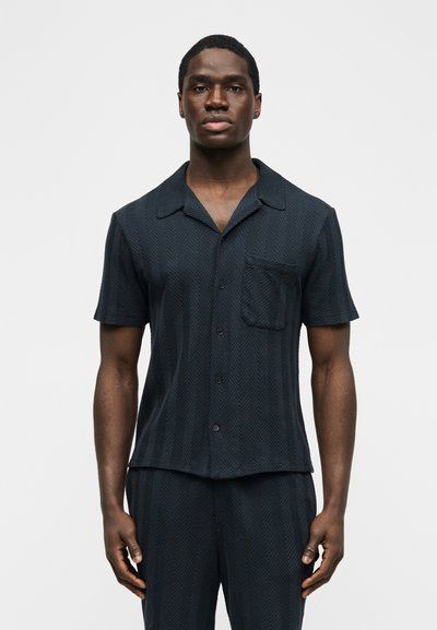 Homme portant une chemise à manches courtes sombre avec poche et un pantalon assorti, debout devant un fond clair uni.