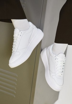Zapatillas blancas de cuero con una textura suave, que cuentan con un cuello acolchado, cierre con cordones y una suela de goma blanca gruesa.