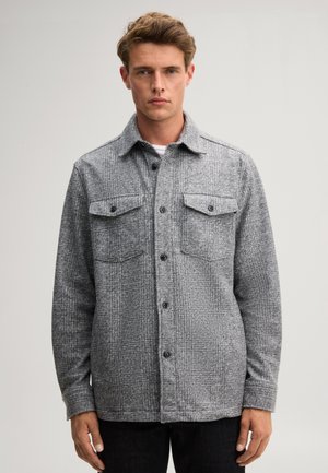 Jonge man die een grijze wollen overshirt met knopen en borstzakken draagt, staand tegen een eenvoudige neutrale achtergrond.