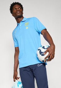 Svetlo modra polo majica z rumenim logotipom Barcelone, v kombinaciji s temno modrimi hlačami in nogometno žogo v rokah. Majica ima ovratnik in kratke rokave.