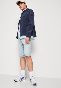 Giacca blu navy strutturata sopra una camicia azzurro chiaro, abbinata a pantaloni corti in denim chiaro e sneakers bianche con dettagli blu.