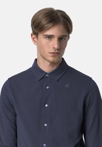 Polo a maniche lunghe blu navy in tessuto texturizzato, con pattina bottonata, colletto a punta e branding discreto sul petto sinistro.