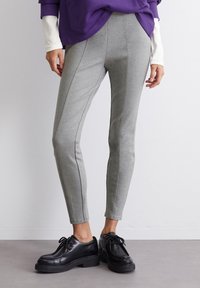 Leggings grigi aderenti realizzati in un tessuto morbido e elastico. Presentano una texture liscia con dettagli a cucitura verticale, abbinati a scarpe nere con lacci.