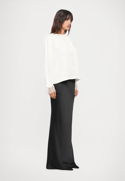 Weiße Bluse mit bindbarem Vorderteil und Spitzensaum an den Ärmeln, kombiniert mit einem glatten schwarzen Maxirock. Schlichte, elegante Silhouette mit weicher Textur.