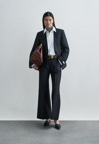 Εξατομικευμένο μαύρο κοστούμι με παντελόνια φαρδιάς γραμμής, λευκό πουκάμισο και γκρι ζιβάγκο. Καφέ δερμάτινη τσάντα, χρυσή αγκράφα ζώνης και μαύρα τακούνια.