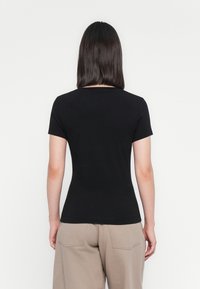 Kvinde med skulderlangt mørkt hår iført en tætsiddende sort kortærmet skjorte og beige bukser, vist fra ryggen mod en hvid baggrund.