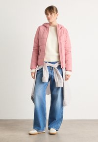 Lichtroze pufferjack met een capuchon, witte gebreide trui, losse blauwe denimbroek en een gestreept shirt om de taille geknoopt. Witte sneakers.