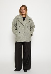 Gai pelēks vilnas sajaukuma dubultā jaku ar lieliem atlokiem un divām priekšējām kabatām, savienots ar melniem plašiem biksēm.