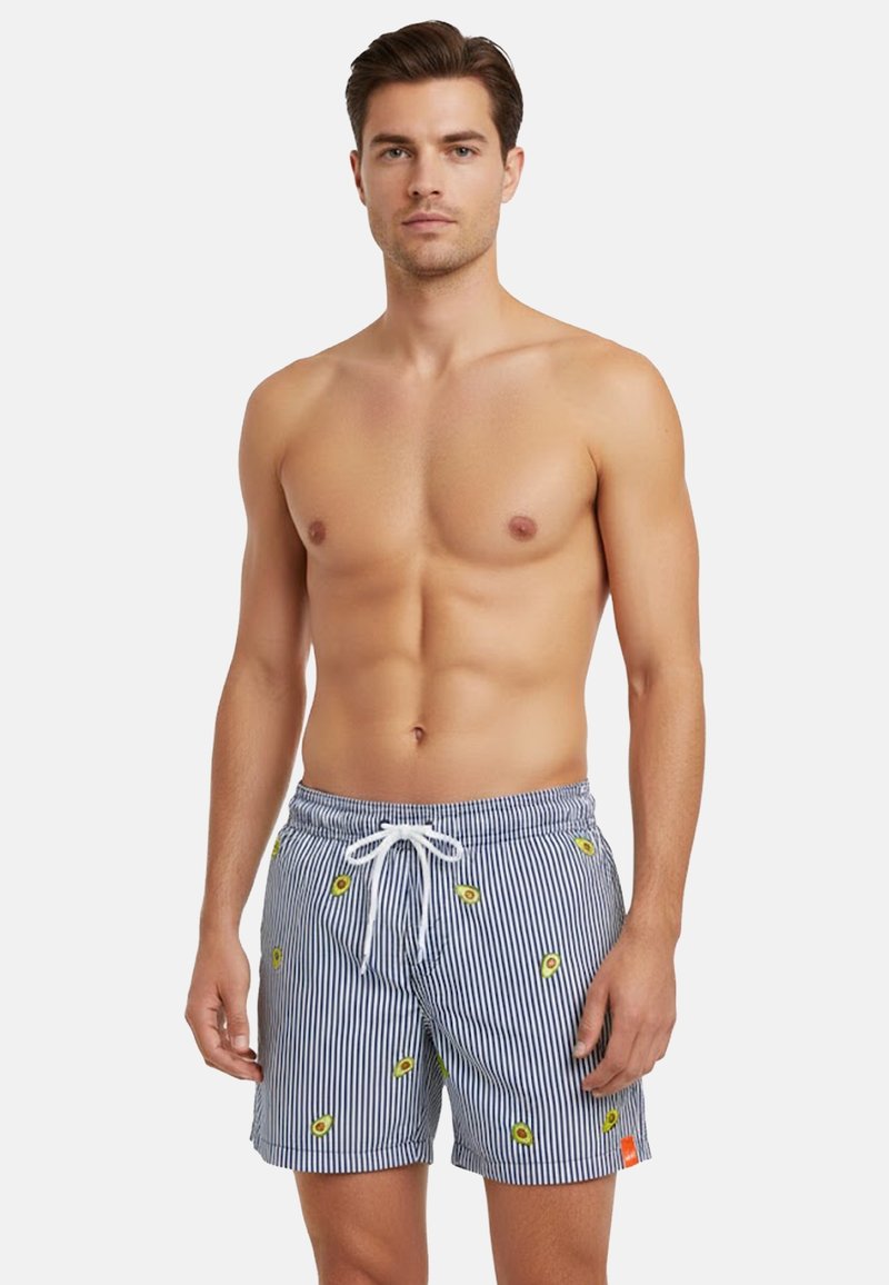 Uomo senza maglietta che indossa pantaloncini da bagno a righe blu e bianche con stampe di avocado, in piedi su uno sfondo bianco uniforme.
