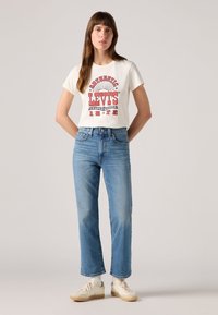 T-shirt en coton blanc avec un graphique rouge de Levi's, associé à un jean droit bleu clair et des baskets crèmes. Design simple, coupe décontractée.