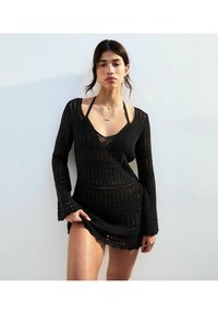 Vestito nero lavorato a maglia con maniche lunghe e scollo a V profondo, caratterizzato da motivi aperti e dettagli testurizzati, che mettono in risalto la pelle sottostante.