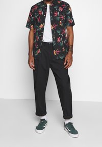 Camicia nera a maniche corte con stampa botanica colorata, T-shirt bianca sotto, pantaloni neri plissettati e sneakers verdi.