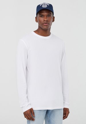 T-shirt bianco a maniche lunghe in cotone, con collo rotondo. Indossato con jeans azzurri chiari e un cappellino blu con logo bianco.