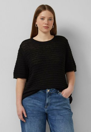 Kobieta o prostych brązowych włosach, nosząca kolczyki koła, czarny teksturowany top z krótkim rękawem i niebieskie dżinsy, stoi z jedną ręką w kieszeni.