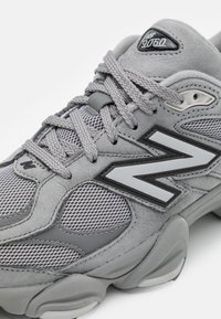 Gros plan d'une basket grise New Balance 9060 avec des panneaux en mesh, des empiècements en suède, des lacets épais et une semelle épaisse.