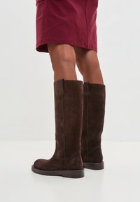 Botas de ante marrón hasta la rodilla con suela plana. Presentan costuras suaves y un diseño de punta redondeada. Tiras de tirón simples y funcionales a cada lado.