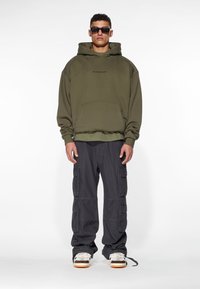 Olive green oversized hoodie with front pocket, paired with dark cargo pants featuring multiple pockets. Model wears sunglasses and sneakers.