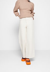 Pantalones crema de pierna ancha combinados con un suéter beige acanalado. Incluye sandalias plataforma naranjas con suela gruesa y tiras ajustables.