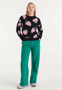 Suéter negro de punto con patrones de fresas rosas y hojas verdes, combinado con pantalones verdes de pierna ancha y zapatillas grises.