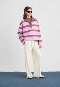 Παντελόνι oversized φούτερ με μοβ και καφέ ρίγες, γιακά και μικρό λογότυπο. Συνδυασμένο με λευκό παντελόνι φαρδιάς γραμμής και γκρίζα παπούτσια τύπου slip-on.