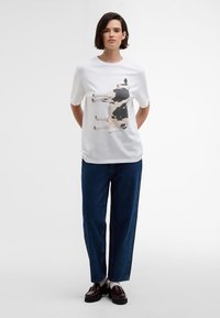 T-shirt branca de algodão com uma grande ilustração de uma vaca em preto e castanho. Combinada com calças de ganga azul largas e mocassins pretos.