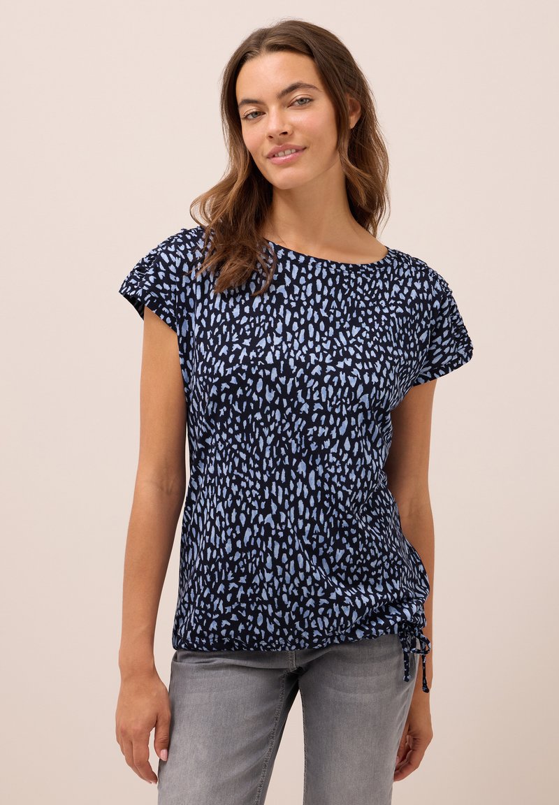 Vrouw in een kort mouw navy top met lichtblauwe abstracte stippen en grijze jeans, staand voor een effen beige achtergrond.