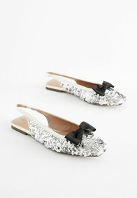 Slingback datar dengan lapisan pereka berwarna perak, hujung runcing, dan aksen reben hitam. Sole berwarna terang, pelapik dalam yang licin.