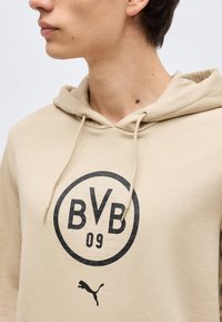 Junge Person in beiger Kapuzenjacke mit BVB 09-Logo und Puma-Logo, seitlich vor einfachem Hintergrund betrachtet.